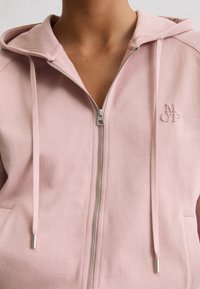 Personne portant un sweat à capuche zippé rose clair avec capuche et cordons, montrant uniquement la zone de la poitrine et du cou.