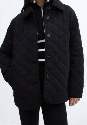 Personen bærer en sort quiltet jakke med knapper over en sort sweater med hvide vandrette striber. Hænderne er delvist i lommerne.