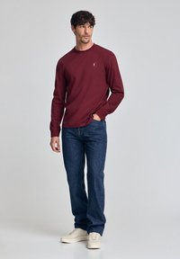 Bordeaux langarmshirt met ronde hals, gecombineerd met donkerblauwe denim jeans en witte sneakers. Heeft een klein logo op de borst.