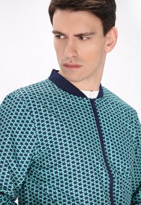 Gevoerde bomberjack met een geometrisch patroon in aqua en marineblauw. Heeft een ronde halslijn en een zachte textuur. Sluit nauw aan.