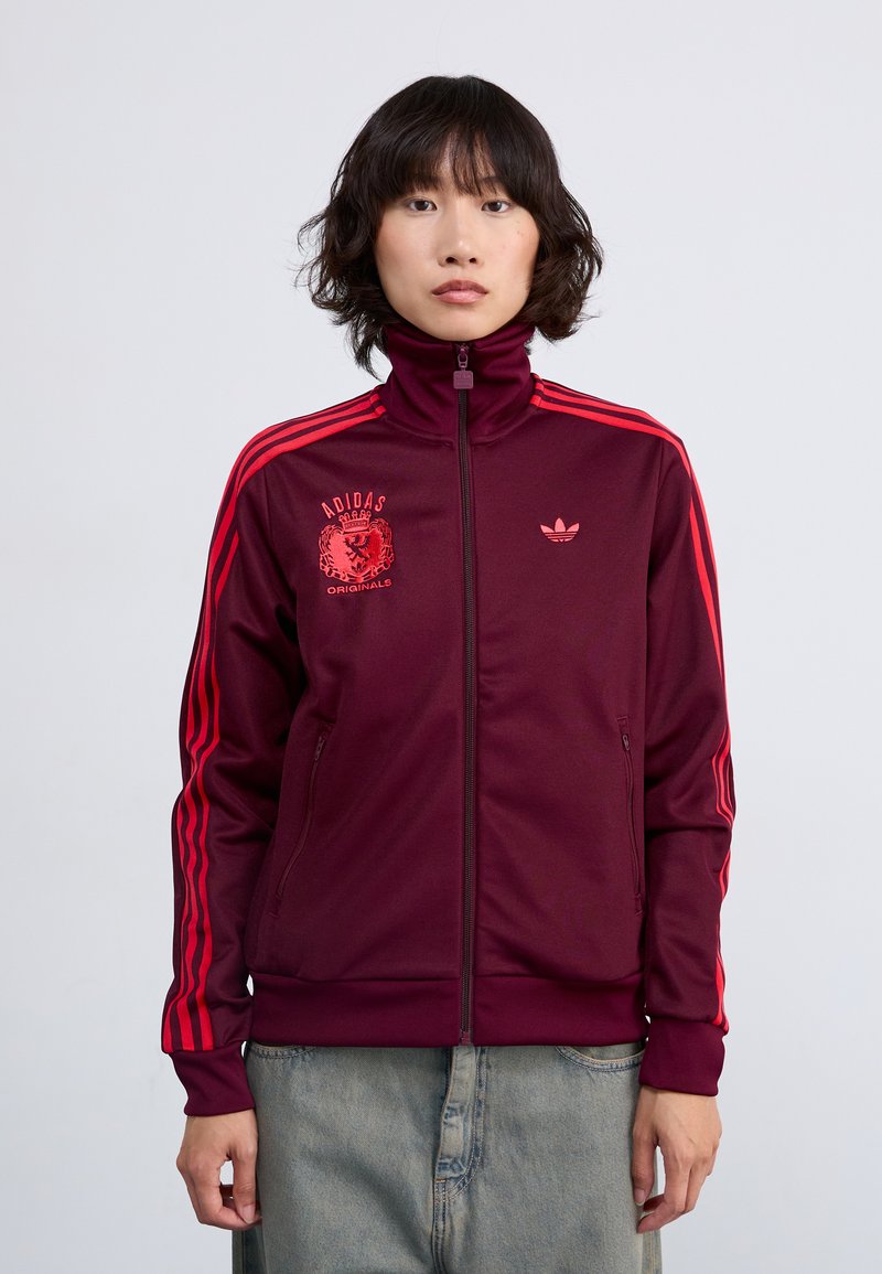Červená maroon adidas sportovní bunda s vysokým límcem, předním zipem a dvěma bočními kapsami. Má červený design s třemi pruhy na rukávech a logem.
