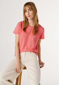 Mujer joven con cabello lacio castaño claro, vestida con una camisa coral de manga corta y pantalones crema, sentada en un taburete de madera contra un fondo blanco.