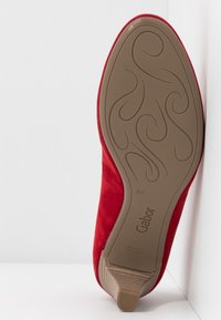 Chaussure en daim rouge avec un design lisse et semi-arqué. Présente une semelle en caoutchouc brun motif et un talon discret. Marque Gabor au dessous.