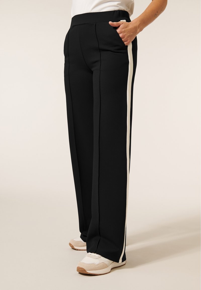 Zwarte sportbroek met een ruime pasvorm, voorzien van crèmekleurige zijstrepen en zakken, gemaakt van een gladde, elastische stof.