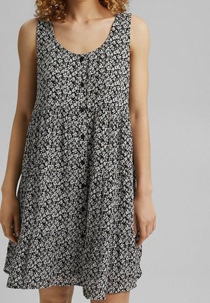 Kobieta w czarnej sukience bez rękawów z małym białym kwiatowym wzorem i guzikami z przodu, na szarym tle.