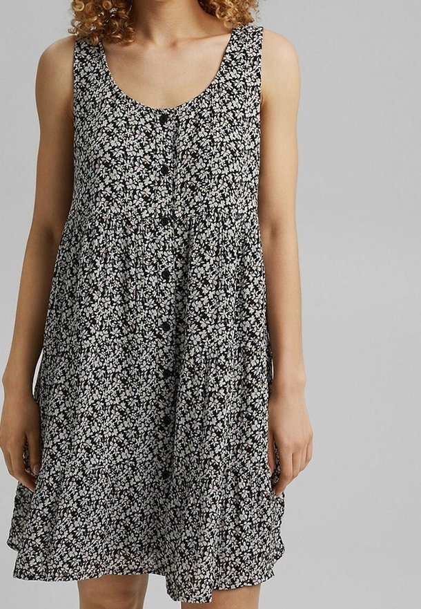 Femme portant une robe noire sans manches avec un petit motif floral blanc et des boutons sur le devant, devant un fond gris.