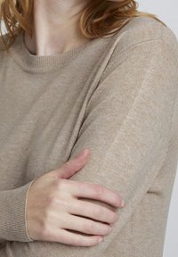 Beiges Strickpullover mit Rundhalsausschnitt, der eine strukturierte Oberfläche und gerippte Bündchen aufweist. Der Stoff wirkt weich und leicht.