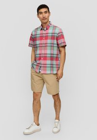 Camicia a maniche corte a quadri nei toni di rosso, verde e blu, con chiusura a bottoni. Abbinata a pantaloni corti beige e sneakers bianche. Vestibilità casual.