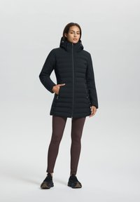 Zwarte pufferjas met een hoge kraag, ritssluiting aan de voorkant en zijzakken, gecombineerd met bruine legging en zwarte sneakers op een neutrale achtergrond.