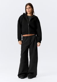 Mustast lühendatud kapuutsiga jakk luku ja esitaskutega, kombineeritud laia säärega mustade pükstega. Lihtne disain, vabaaja kangas ja mugav lõige.