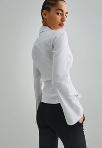 Bílá halenka s dlouhým rukávem a úzkým pasem, s knoflíkovým detailem a rozšířenými rukávy, v kombinaci s černými kalhotami. Texturovaná tkanina s hladkým povrchem.
