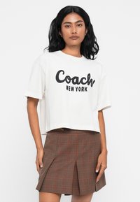 Camiseta corta blanca con el texto "Coach New York" en negro, combinada con una falda de cuadros marrón con acentos rojos y plisados.