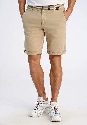 Homme portant un short beige retroussé avec une ceinture tressée et des chaussures de sport blanches, debout avec les mains dans les poches.