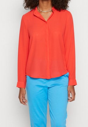 Vrouw die een felrode blouse met lange mouwen, blauwe broek en een zilveren kettinkje draagt, staand tegen een effen achtergrond.