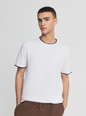 Giovane uomo che indossa una maglietta bianca con rifiniture nere e pantaloni marroni, in piedi davanti a uno sfondo grigio chiaro uniforme.