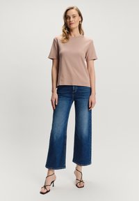 Kobieta stojąca w jasnobrązowej bluzce z krótkim rękawem, niebieskich szerokich dżinsach, czarnych sandałach na obcasie i złotych kolczykach na jednolitym tle.