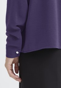 Vijolična srajca z dolgimi rokavi in gumbom na manžetah, mehke teksture in ravnim robom, v kombinaciji sčrnimi hlačami, z roko, ki počiva ob strani.