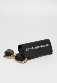 Ochelari de soare cu lentile întunecate și brațe din tortoiseshell cu accente aurii, așezați lângă un etui negru pe care este imprimat cu alb "RETROSUPERFUTURE".