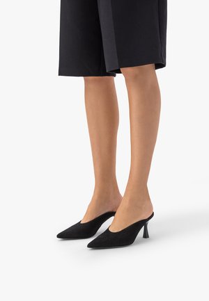 Nogi w czarnych, sięgających połowy uda szortach i czarnych, spiczastych mule na cienkich wysokich obcasach na białym tle.