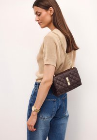 Borsa a tracolla marrone trapuntata con catena dorata, dotata di patta frontale e chiusura, abbinata a jeans a vita alta blu chiaro e una top beige.