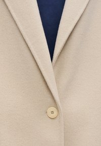 Beige wool blend coat featuring peaked lapels, a single creamy button closure, and a smooth texture. Underneath is a dark navy fabric.