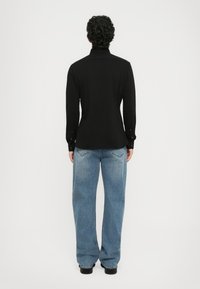Person med krøllete hår iført en langermet svart skjorte og løse blå jeans, stående og vendt mot en ensfarget hvit bakgrunn.