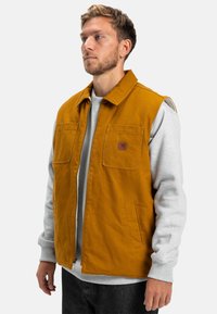 Gilet senza maniche di colore senape con due tasche sul petto e una tasca laterale, realizzato in un tessuto resistente, indossato sopra un maglione grigio.