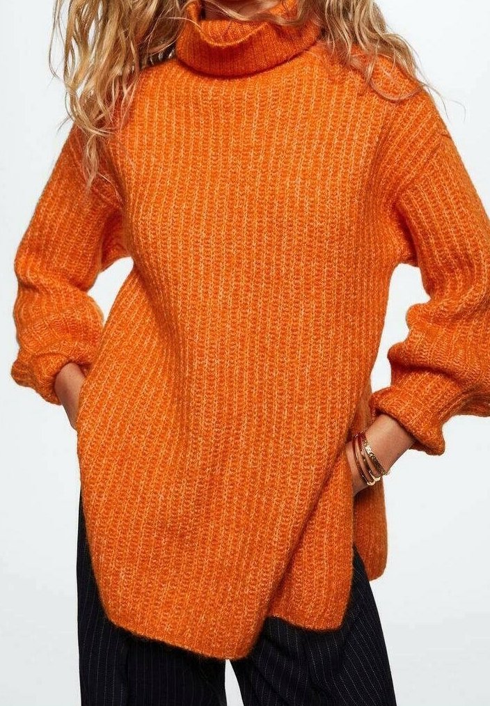 Pomarańczowy sweter z golfem, wykonany na drutach, o prążkowanej fakturze, w luźnym kroju z bocznymi rozcięciami. Zestawiony z ciemnymi spodniami w prążki.
