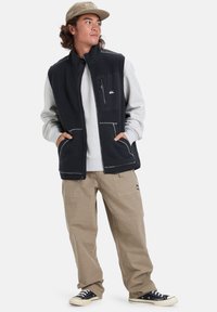 Black fleece vest with gray sleeves, zippered chest pocket, and side pockets, paired with khaki pants and black sneakers.