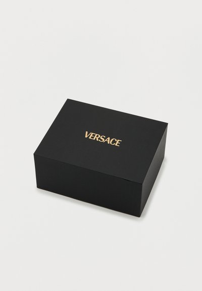 Μαύρο ορθογώνιο κουτί με υφή στην επιφάνεια, που φέρει τη λέξη "VERSACE" με χρυσά ανάγλυφα γράμματα στην κορυφή.