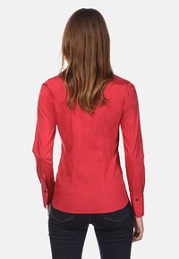Rode getailleerde blouse met lange mouwen, met knopen bij de manchetten en een gladde textuur, contoureer naden die de vorm benadrukken, gedragen met donkere jeans.
