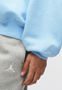 Detailný záber ruky odpočívajúcej pri páse, oblečenej v modrom mikinovom svetri a šedých teplákoch, oboje s logom Jumpman.