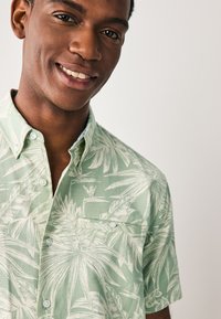 Camicia a maniche corte verde chiaro con bottoni e motivo testurizzato, decorata con disegni di foglie di palma bianche. Presenta una tasca sul petto e un colletto.