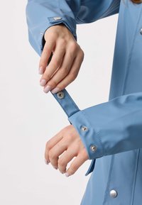 Ljusblå regnjacka med en slät yta, utrustad med silverfärgade tryckknappar vid muddarna. Muddarna är justerbara och åtsittande.