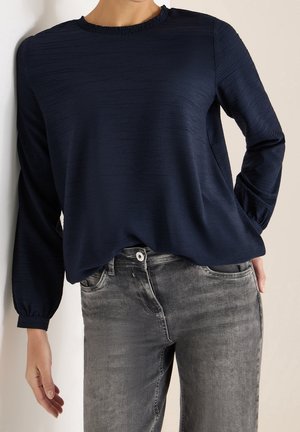 Femme portant un chemisier bleu marine texturé à manches longues avec un col volanté et un jean gris délavé, posant avec une main dans la poche.