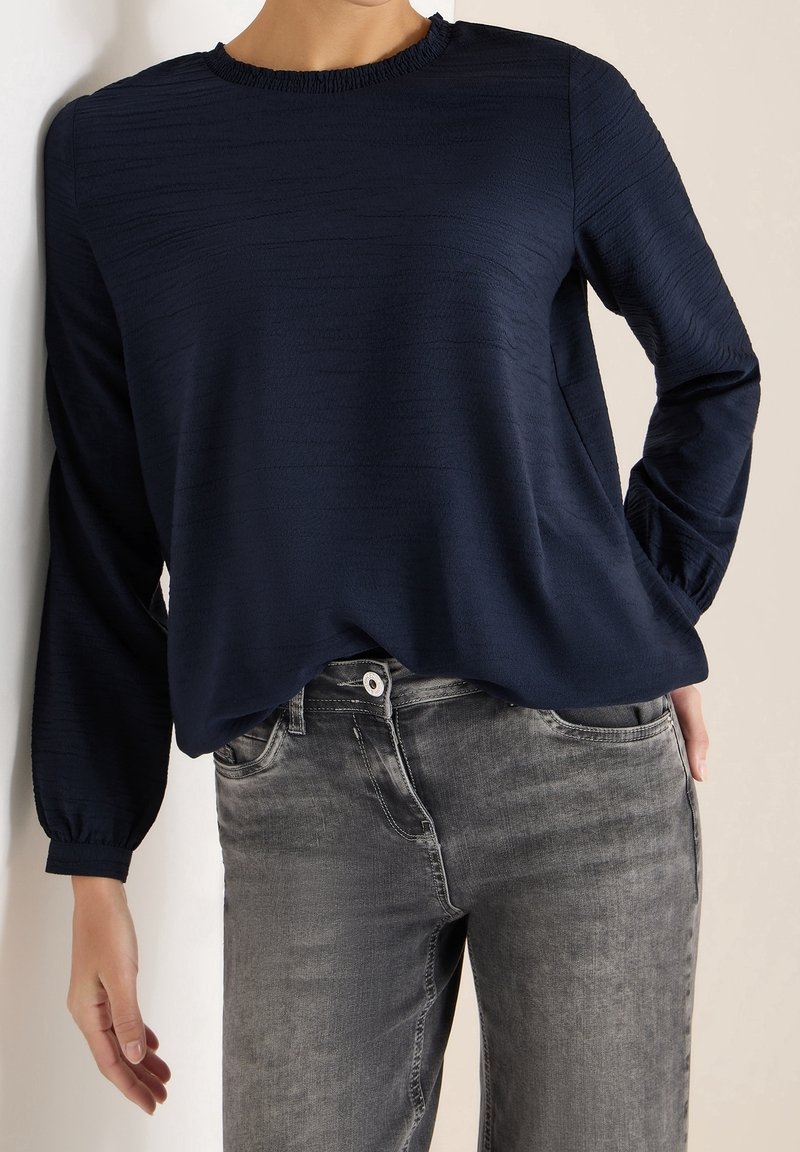 Femme portant un chemisier bleu marine texturé à manches longues avec un col volanté et un jean gris délavé, posant avec une main dans la poche.