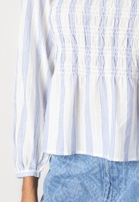 Chemisier blanc à rayures verticales bleues, corsage froncé, coupe décontractée et manches longues volumineuses, associé à un bas en denim bleu clair.