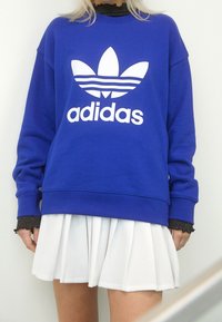 En person som bär en blå Adidas-tröja med vitt logotyp, en svart långärmad topp under, och en vit plisserad kjol som står mot en vit bakgrund.