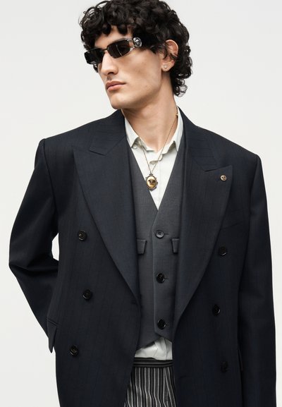 Άνδρας με σγουρά σκούρα μαλλιά που φοράει μαύρο διπλόμπουκο σακάκι με ρίγες, γκρι γιλέκο, ανοιχτό πουκάμισο, χρυσό μενταγιόν, γυαλιά ηλίου και ριγωτό παντελόνι.