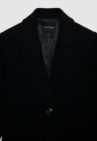 Cappotto in pura lana nera con rever a notched, chiusura frontale con un solo bottone e due tasche frontali. L'interno è foderato con un tessuto a contrasto. Etichetta della taglia S visibile.