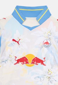 Camisa de Puma con un ligero estampado floral en colores pastel. Presenta un cuello azul con acentos amarillos y un logo de Red Bull en rojo. Tela liviana.