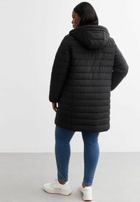 Abrigo acolchado negro con capucha, de corte midi, con costuras horizontales. Lleva puesto sobre unos vaqueros azules y zapatillas blancas.