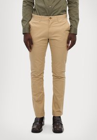 Bežna bombažna hlače s slim fit krojem, z gumbom za zapiranje, zankami za pas in žepi. Oblečene z temnimi čevlji in zeleno srajco.