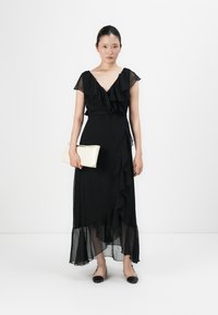 Frau, die ein schwarzes, rüschendes Midikleid trägt, eine beige Clutch hält und schwarze sowie beige Ballerinas trägt, vor einem weißen Hintergrund steht.