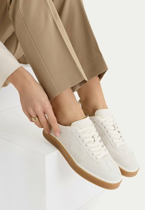 Osoba ve světle hnědých kalhotách si sedí na bílém bloku a nazouvá bílé sportovní boty se šněrováním a hnědými podrážkami.