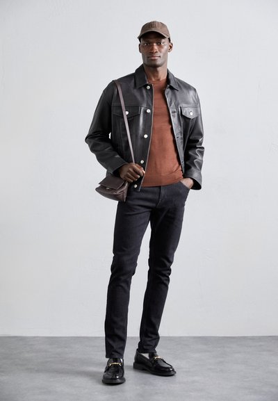 Black leather jacket, rust sweater, dark jeans, and black loafers with gold accents. Brown cap and brown shoulder bag. Simple, modern design.
