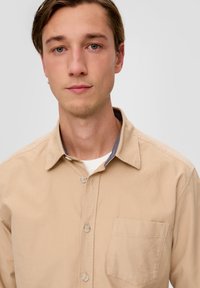 Beige Hemd mit Knopfleiste und Kragen, ausgestattet mit einer einzelnen Brusttasche und einem kontrastierenden Innenkragen. Hergestellt aus strukturiertem Baumwollstoff.