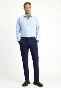 Camicia button-up azzurro chiaro con taschino sul petto, abbinata a pantaloni blu scuro e scarpe nere, in piedi su uno sfondo neutro.
