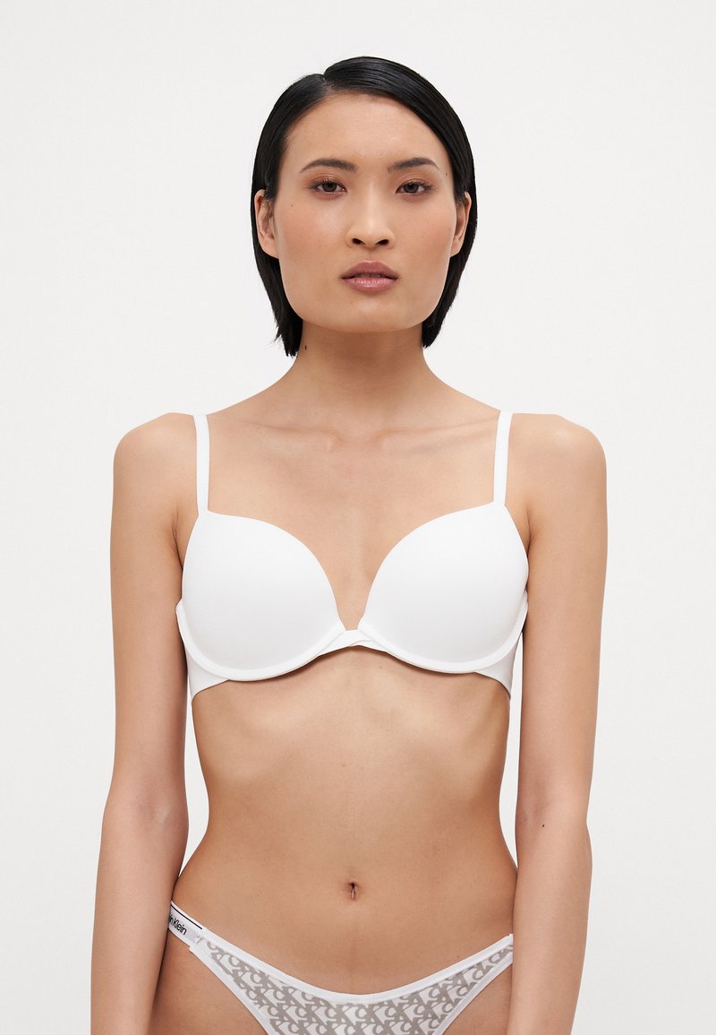 White padded bra with adjustable straps, featuring smooth fabric and a deep V-neckline. Paired with white sheer underwear with logos.