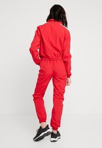 Röd jacka och byxor träningsdräkt med vita sidodetaljer, elastisk midja och smalnande ärmslut. Lättviktig, slät textur. Svarta sneakers synliga.